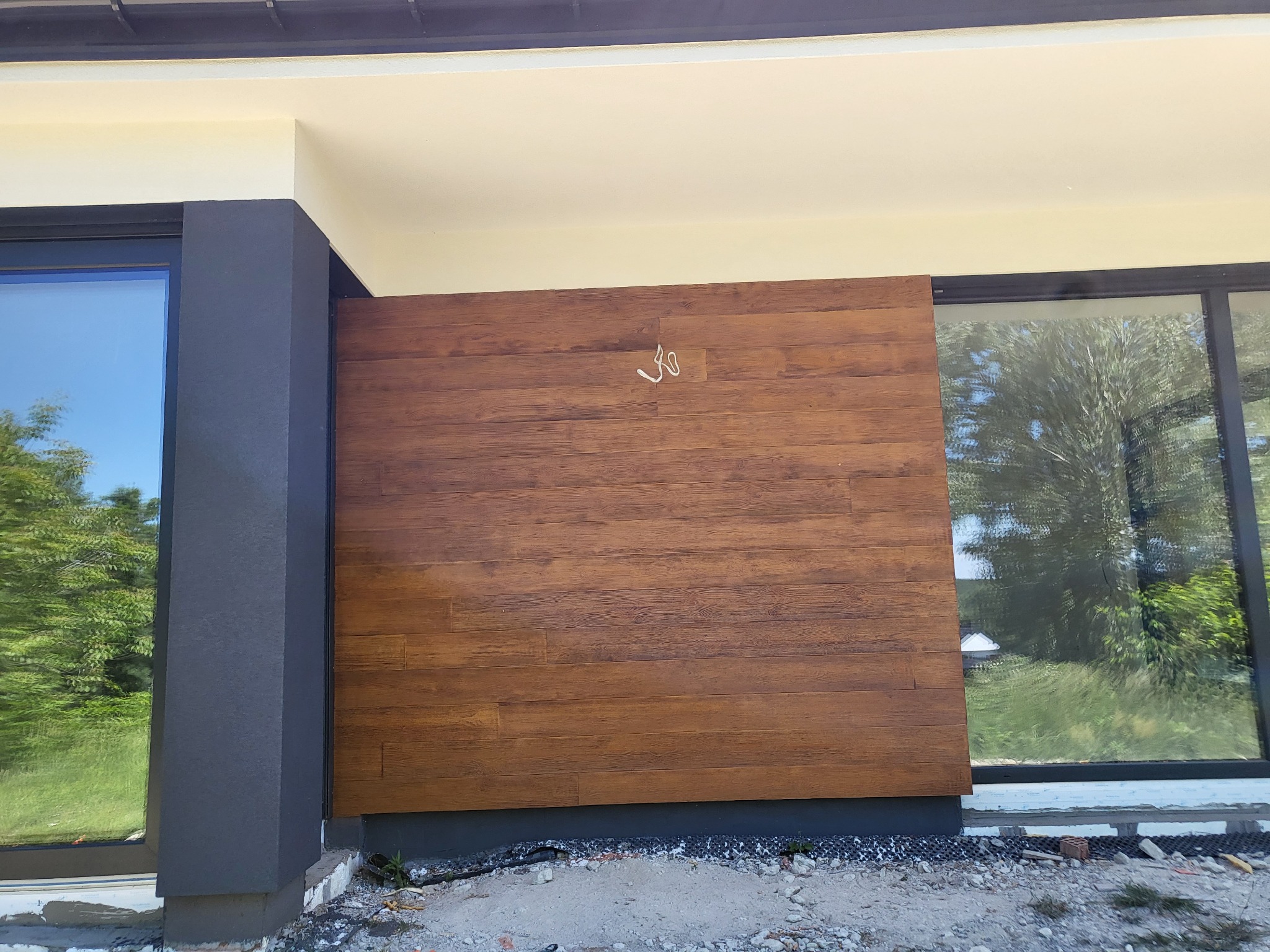 Nowoczesna elewacja domu z drewnopodobną okładziną i dużymi oknami, wykończona szarą kolumną. Widoczne detale wykończeniowe i fragment otoczenia.