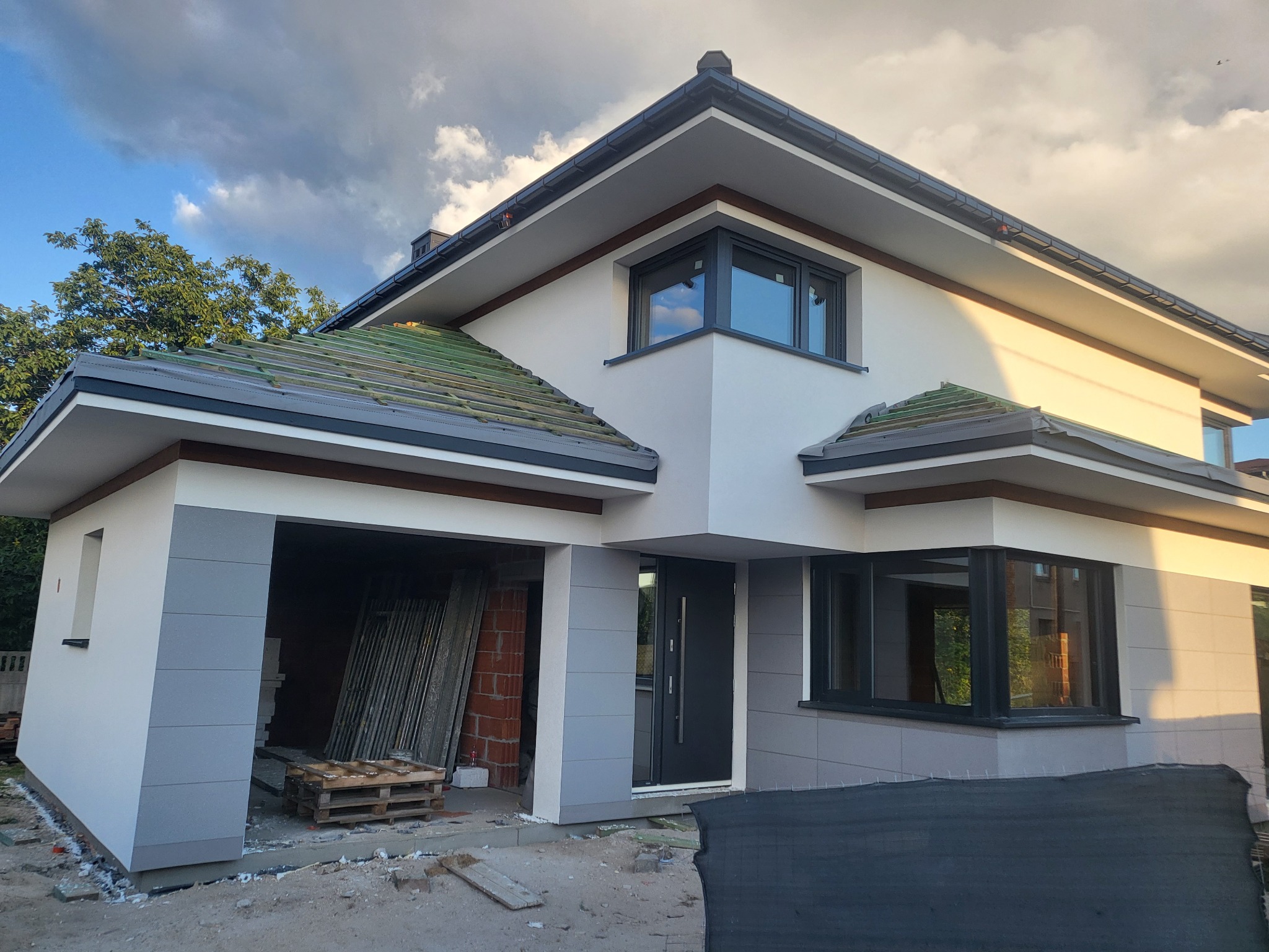 Nowoczesny dom jednorodzinny z elewacją w odcieniach bieli i szarości, z widocznym garażem w trakcie wykończenia. Częściowo położone dachówki.