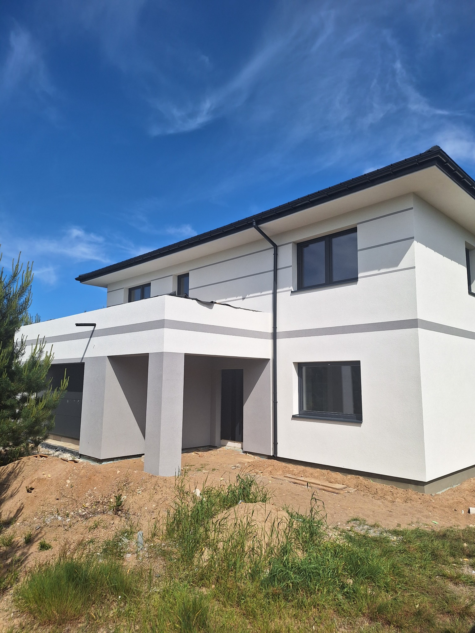 Dwukondygnacyjny dom z białą elewacją i szarymi akcentami, nowoczesna architektura, błękitne niebo w tle, widok z poziomu gruntu, teren wokół domu jeszcze niezagospodarowany.