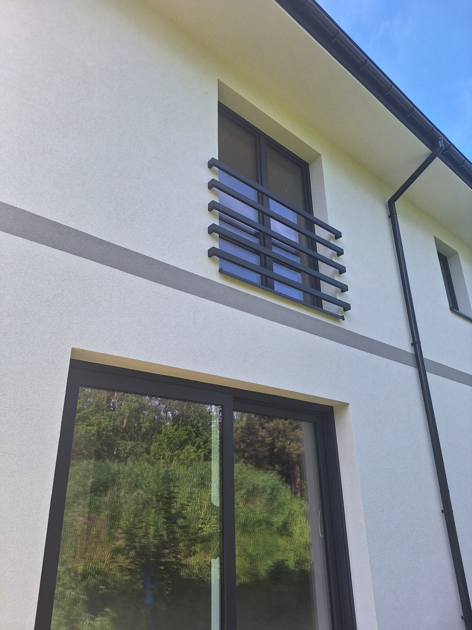 Biała elewacja domu z szarym pasem, czarnymi oknami i balkonem. Widok z dołu ukazuje fakturę tynku i detale architektoniczne. Błękitne niebo w tle.