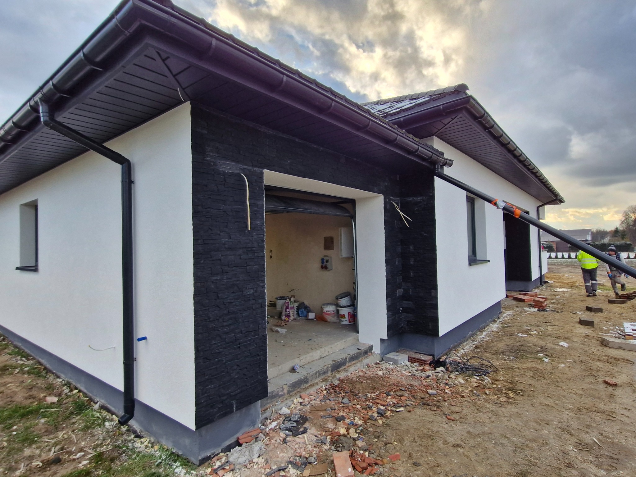 Dwukondygnacyjny budynek z białą elewacją i czarnymi elementami dekoracyjnymi, w trakcie prac wykończeniowych. Widoczne materiały budowlane i pracownicy.