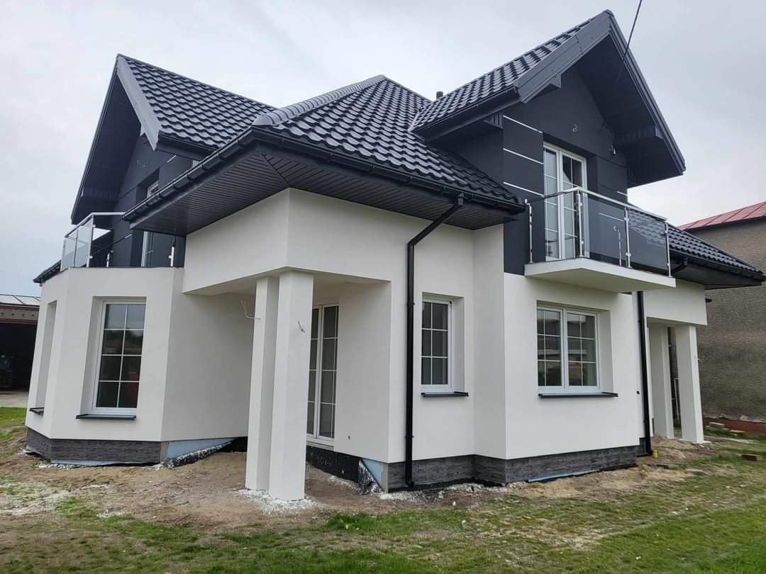 Nowoczesny dom z białą elewacją i czarnym dachem, widoczne balkony z szklanymi barierkami. Minimalistyczny design, świeżo wykończony, zieleń wokół.