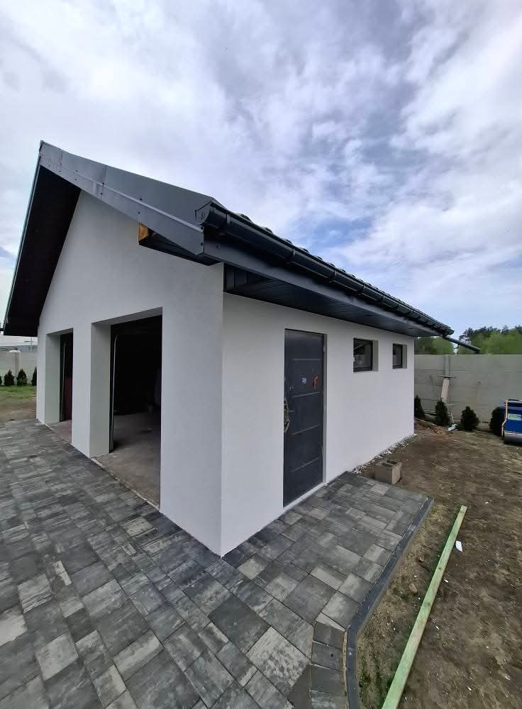Biały budynek gospodarczy z ciemnym dachem i drzwiami, świeżo po tynkowaniu. Widoczne detale wykończenia i kostka brukowa wokół budynku.