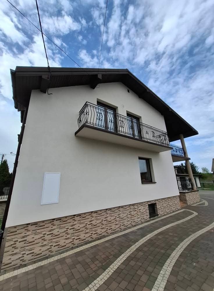 Elewacja domu z balkonem i dekoracyjnym wykończeniem. Jasna ściana kontrastuje z ciemnym dachem i balustradami. Widoczny fragment chodnika z kostki.