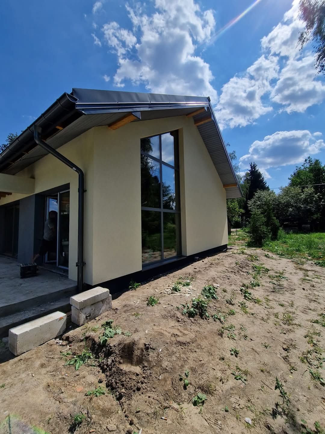 Jasna elewacja domu z dużym oknem i szarą stolarką, widok z zewnątrz w słoneczny dzień. Elementy wykończeniowe przy dachu. Teren wokół domu w trakcie prac ziemnych.