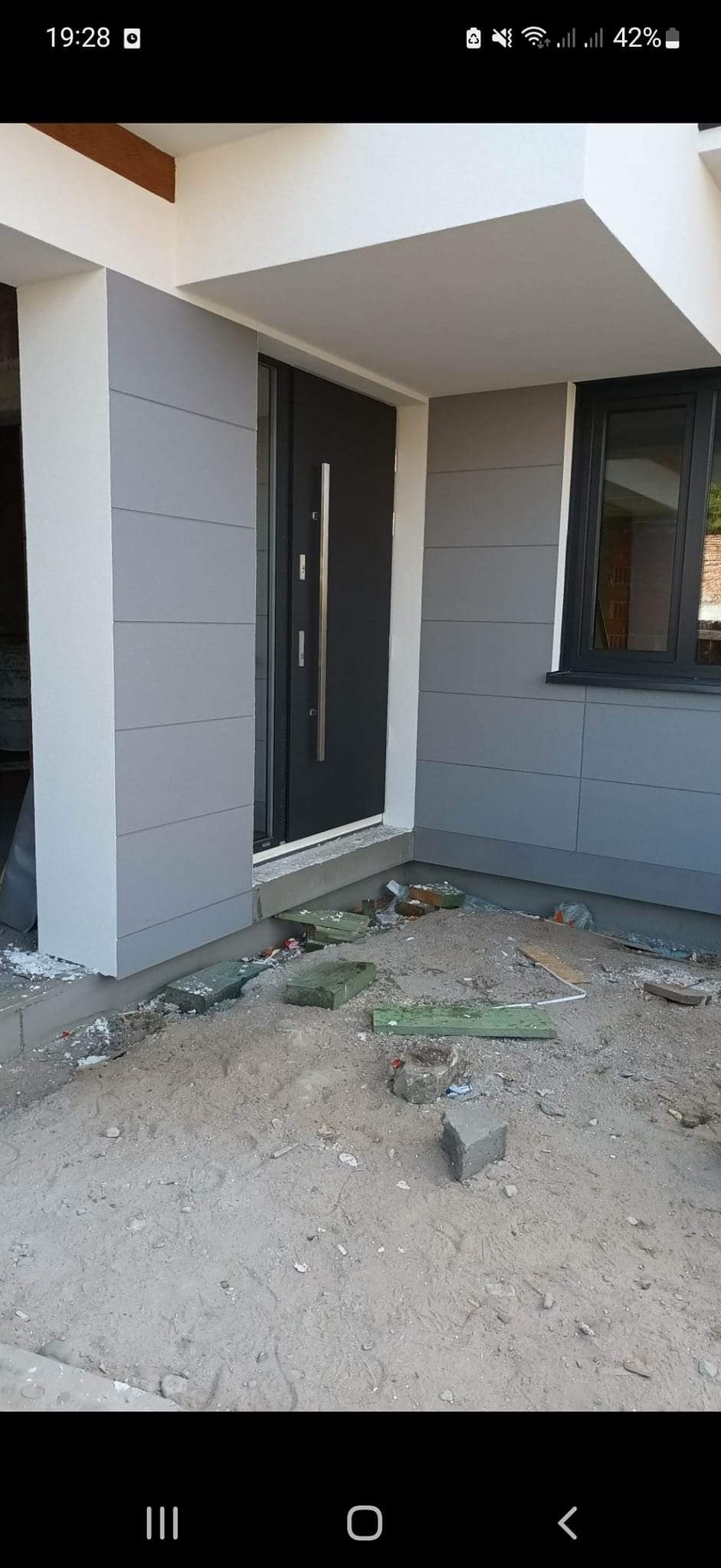 Wykończona elewacja domu z szarymi panelami, czarnymi drzwiami i oknem. Widoczne pozostałości po pracach budowlanych na gruncie przed budynkiem. Ujęcie z bliska.