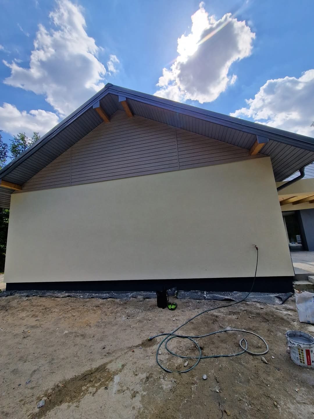 Elewacja domu po tynkowaniu, z widoczną strukturą tynku i pomalowanym cokołem. W tle błękitne niebo z chmurami, na pierwszym planie wąż i wiadro po farbie.