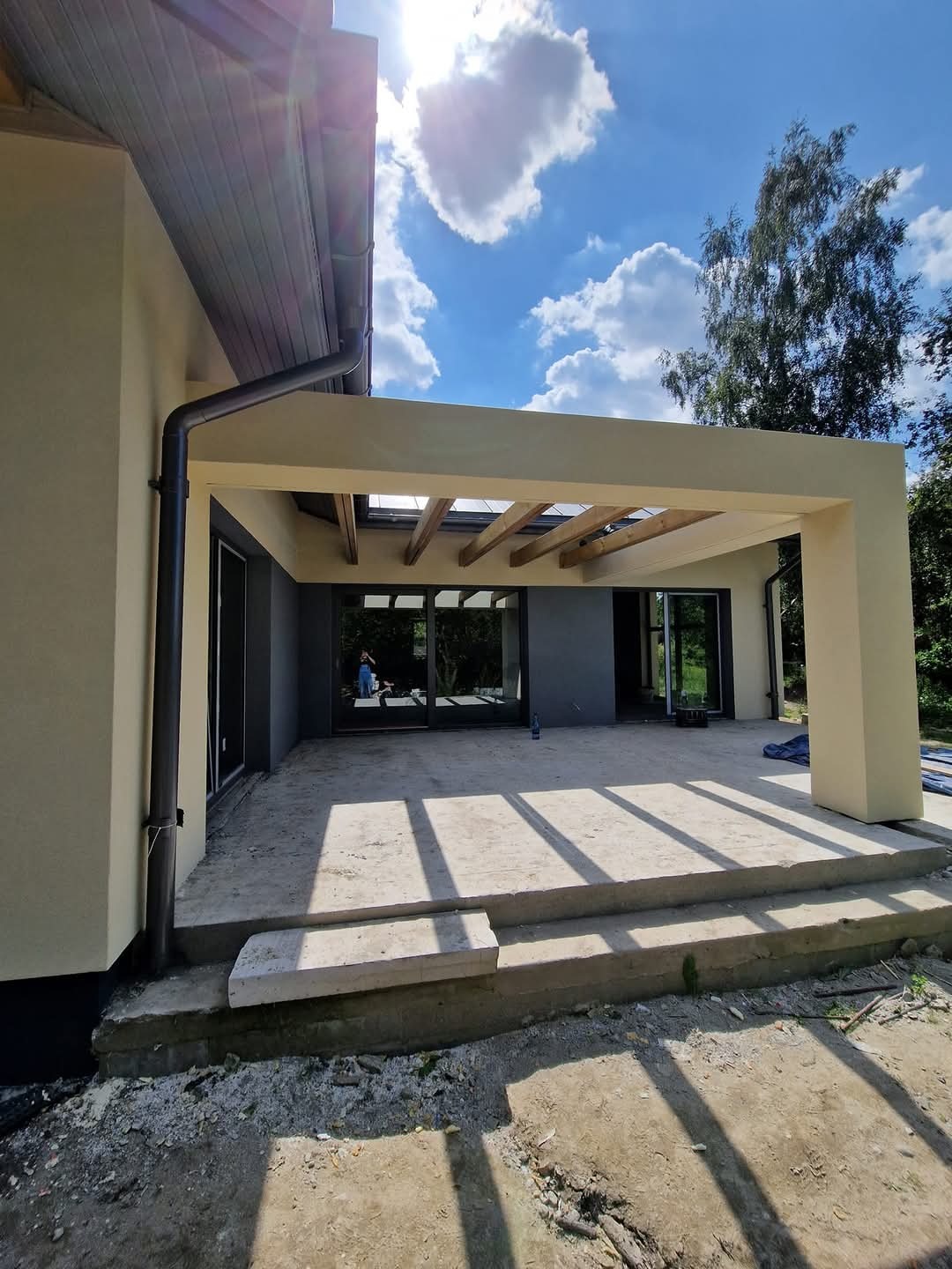 Jasna elewacja domu z zadaszonym tarasem w trakcie budowy, widoczne drewniane elementy konstrukcyjne i betonowe schody, błękitne niebo w tle.