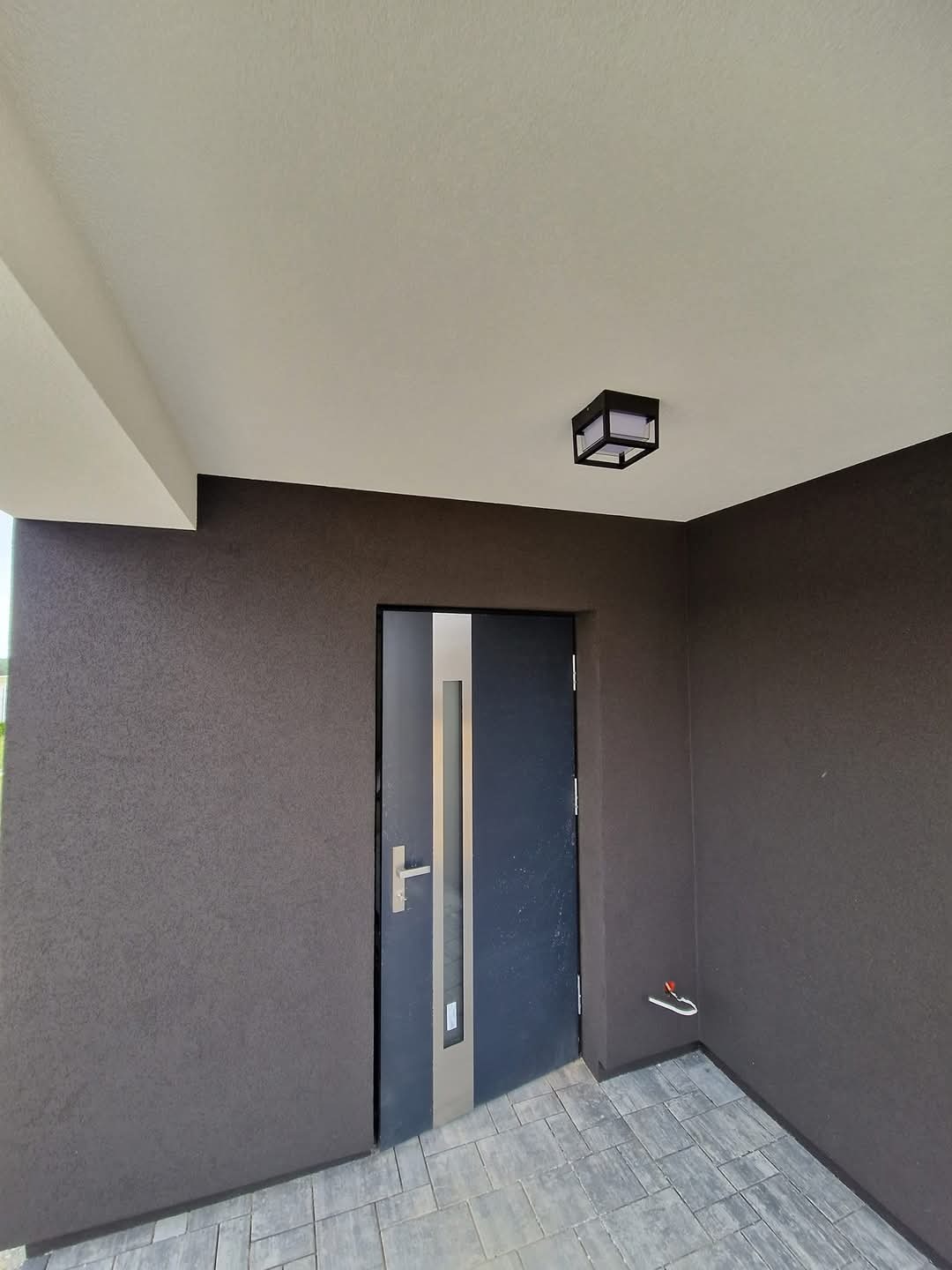 Eleganckie wejście do domu z ciemną elewacją o wyraźnej fakturze, nowoczesnymi drzwiami z elementami stali nierdzewnej i kostką brukową. Oświetlenie sufitowe.