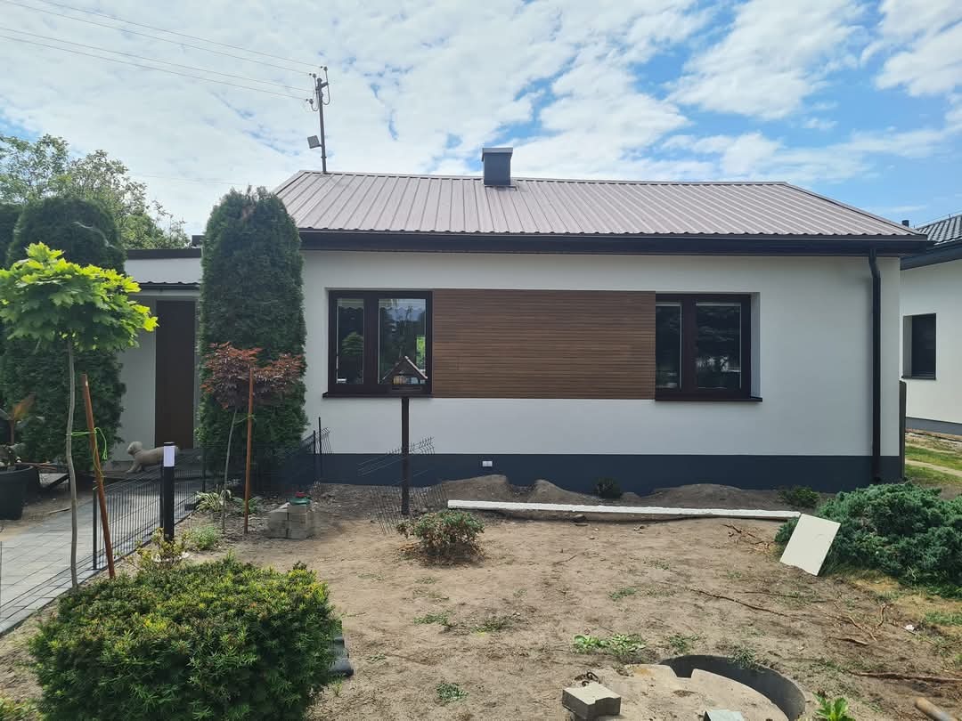 Biały dom z brązowym dachem i drewnianą elewacją, widok z zewnątrz, zieleń w ogrodzie, błękitne niebo z chmurami. Nowoczesne wykończenie.