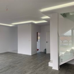 MUCKENS-BUD Firma Ogólno-Budowlana Kompleksowe wykończenie - Wnętrze po remoncie: białe ściany, szara podłoga imitująca drewno, oświetlenie LED w suficie podwieszanym, szklana balustrada schodów, widok na korytarz z pomalowanymi drzwiami w tle.