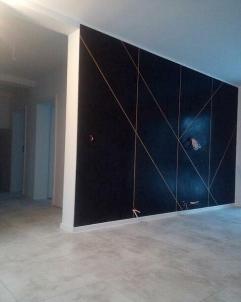 Ściana pomalowana na czarno z geometrycznym wzorem z cienkich, złotych linii, widoczne wystające kable oraz szare płytki podłogowe w pustym pomieszczeniu.