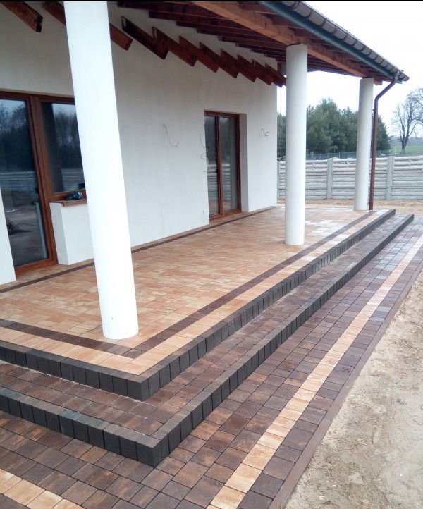 Wykończony taras z kostki brukowej w dwóch odcieniach brązu, z trzema stopniami i kolumnami podtrzymującymi zadaszenie, widok z boku.