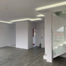 Wnętrze po remoncie: białe ściany, szara podłoga imitująca drewno, oświetlenie LED w suficie podwieszanym, szklana balustrada schodów, widok na korytarz z pomalowanymi drzwiami w tle.