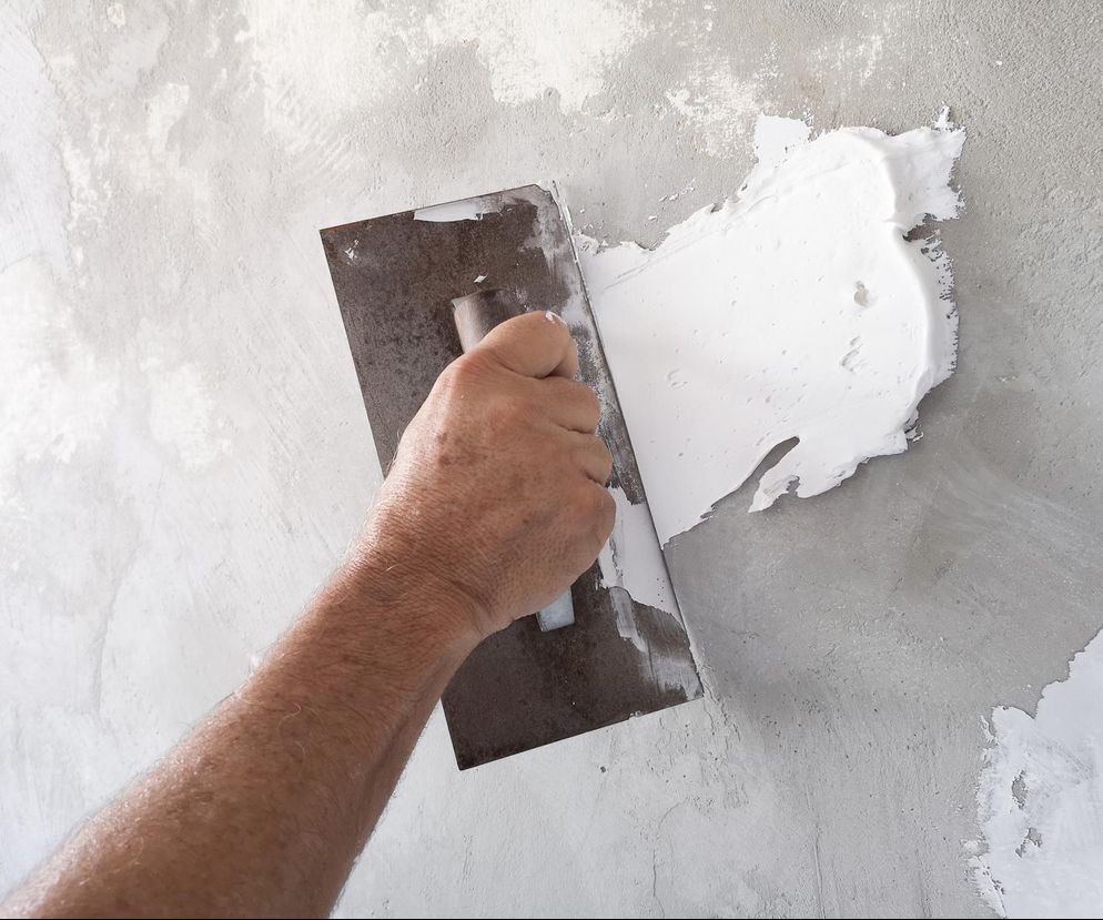 Nakładanie gładzi szpachlowej na ścianę za pomocą metalowej szpachelki, widoczne nierówności i struktura surowej ściany.