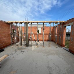 Zakład Usług Budowlanych Jan Tomala - Wnętrze budowanego domu z czerwonej cegły, widoczne metalowe podpory, drewniane belki stropowe, betonowa wylewka podłogowa i fragment drabiny oparty o ścianę.