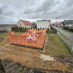 Wznoszone ściany parterowego domu z czerwonej cegły ceramicznej, zabezpieczone czarną folią hydroizolacyjną, na tle wiejskiej zabudowy w pochmurny dzień.