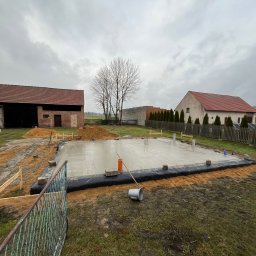 Zakład Usług Budowlanych Jan Tomala - Świeżo wylana płyta fundamentowa na budowie domu jednorodzinnego, zabezpieczona czarną folią, widoczne szalunki i przygotowane zbrojenie, w tle budynki gospodarcze i drzewo bez liści, pochmurne niebo.