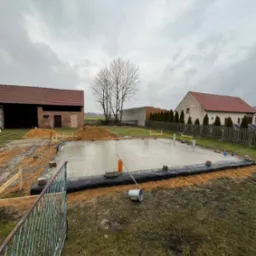 Świeżo wylana płyta fundamentowa na budowie domu jednorodzinnego, zabezpieczona czarną folią, widoczne szalunki i przygotowane zbrojenie, w tle budynki gospodarcze i drzewo bez liści, pochmurne niebo.
