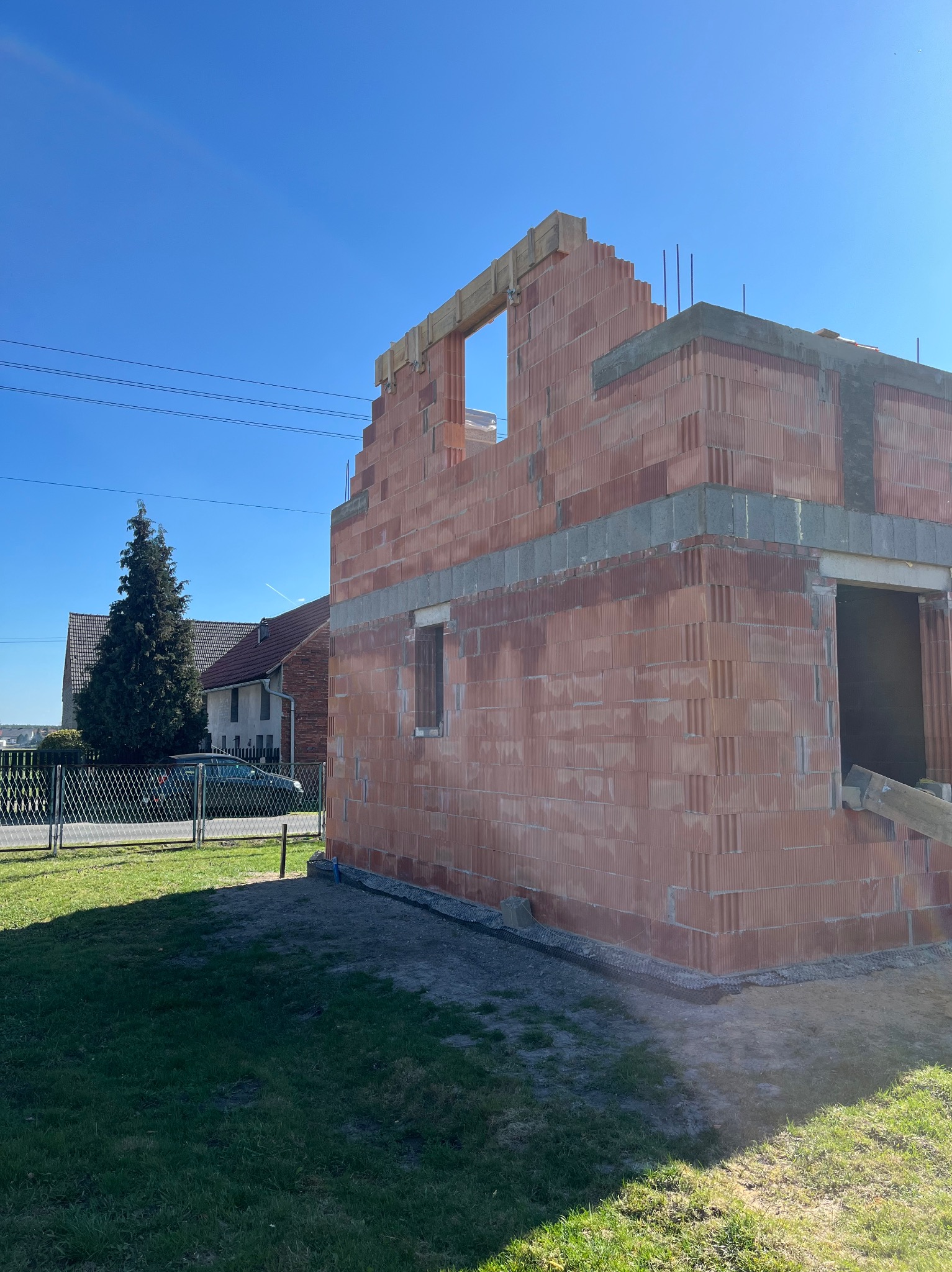 Niedokończony budynek z czerwonej cegły ceramicznej z widocznym zbrojeniem, w tle dom jednorodzinny i wysokie drzewo iglaste pod błękitnym niebem.