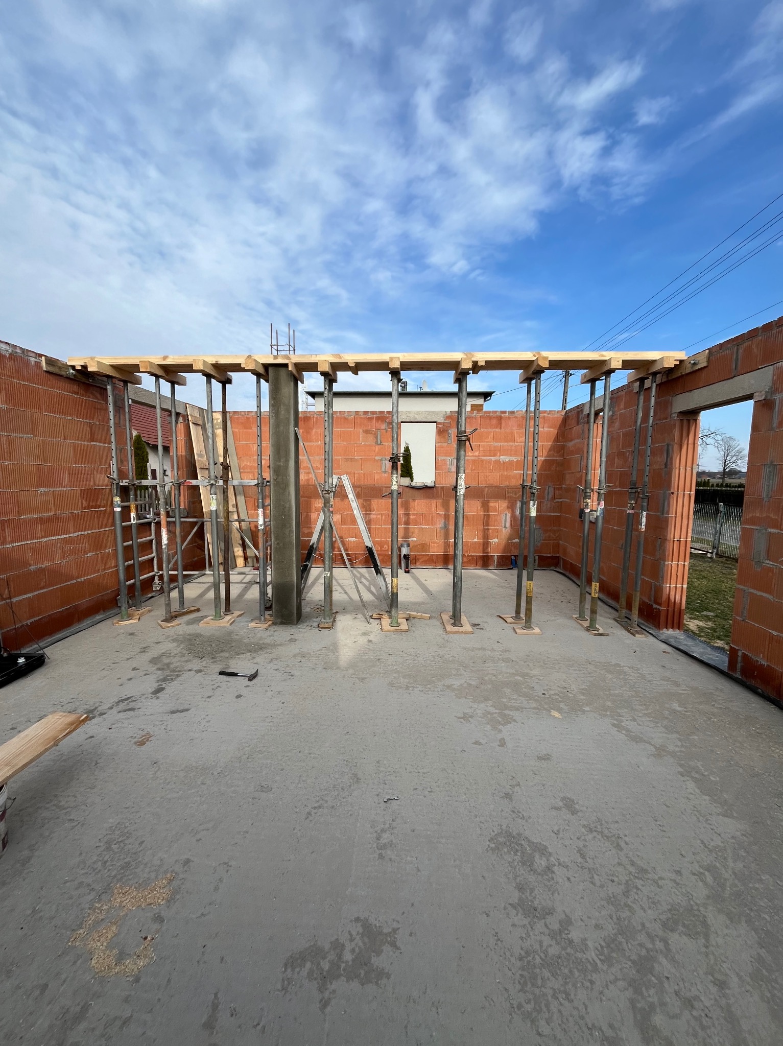 Wnętrze budowanego domu z czerwonej cegły, widoczne metalowe podpory, drewniane belki stropowe, betonowa wylewka podłogowa i fragment drabiny oparty o ścianę.