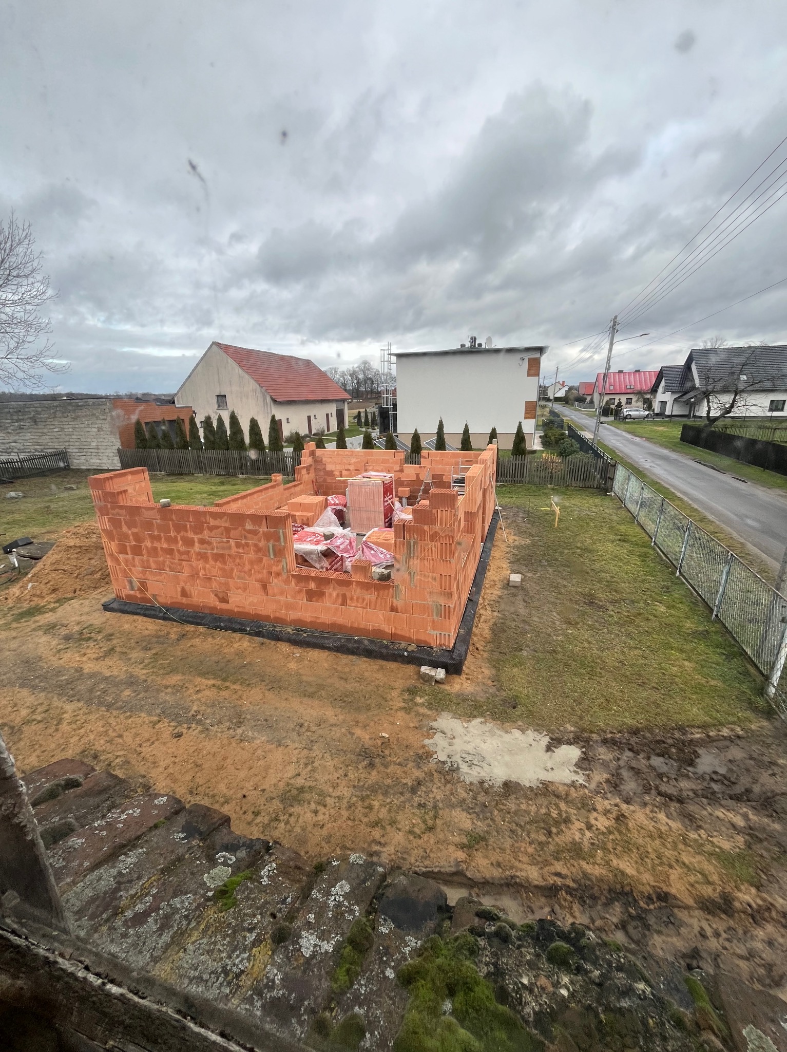 Wznoszone ściany parterowego domu z czerwonej cegły ceramicznej, zabezpieczone czarną folią hydroizolacyjną, na tle wiejskiej zabudowy w pochmurny dzień.