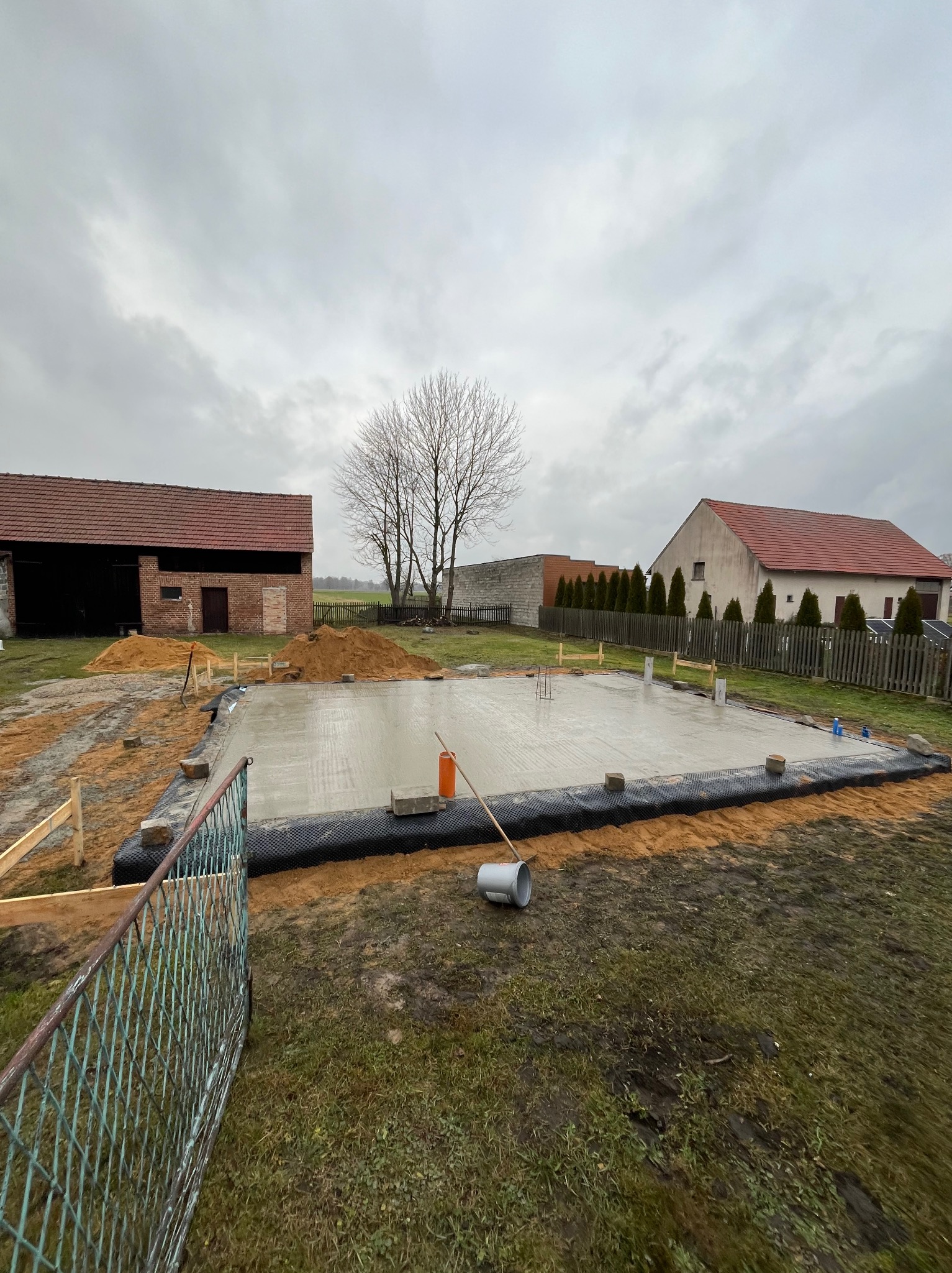 Świeżo wylana płyta fundamentowa na budowie domu jednorodzinnego, zabezpieczona czarną folią, widoczne szalunki i przygotowane zbrojenie, w tle budynki gospodarcze i drzewo bez liści, pochmurne niebo.