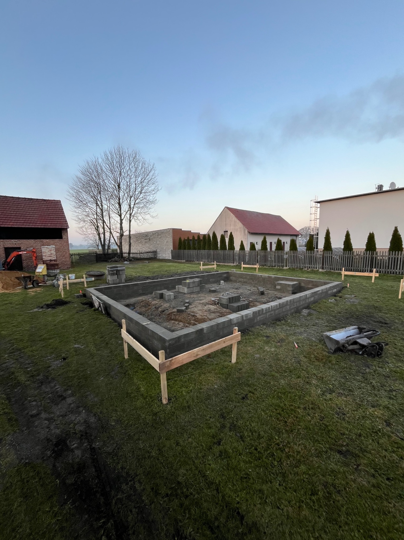 Wylane fundamenty z bloczków betonowych na trawiastym terenie budowy, widoczny wykop i szalunki, w tle budynki gospodarcze i koparka.