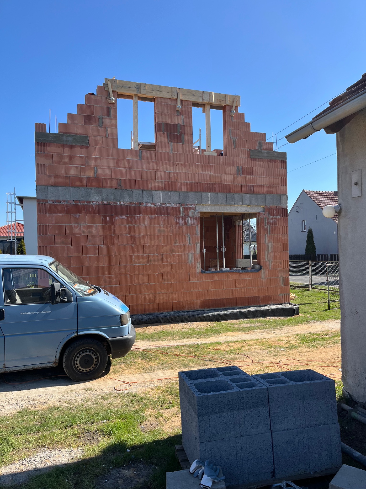 Wznoszenie dwukondygnacyjnego domu z czerwonej cegły ceramicznej, widoczna konstrukcja okien i drewniane nadproża, niebieski bus budowlany na pierwszym planie, stos szarych bloczków betonowych obok.