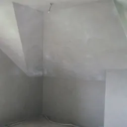 Wnętrze poddasza po nałożeniu szarego tynku wapienno-cementowego, widoczne kable elektryczne przy podłodze i surowa żarówka.