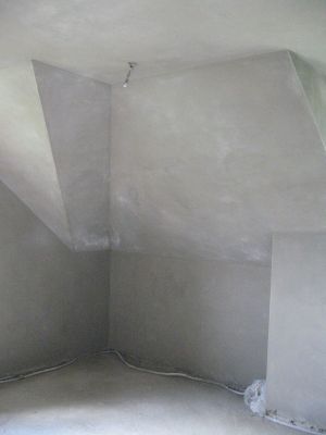 Wnętrze poddasza po nałożeniu szarego tynku wapienno-cementowego, widoczne kable elektryczne przy podłodze i surowa żarówka.