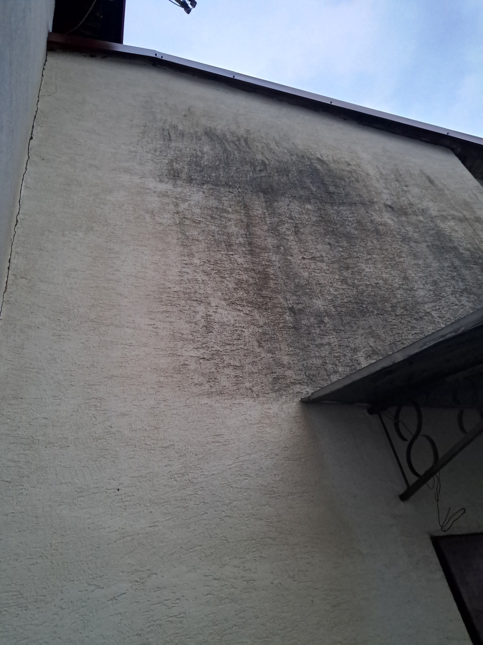 Zewnętrzna ściana budynku z widocznym silnym zabrudzeniem i zaciekami w górnej części, kontrastującym z czystszą dolną partią, pod kątem, ukazująca potrzebę odświeżenia elewacji.