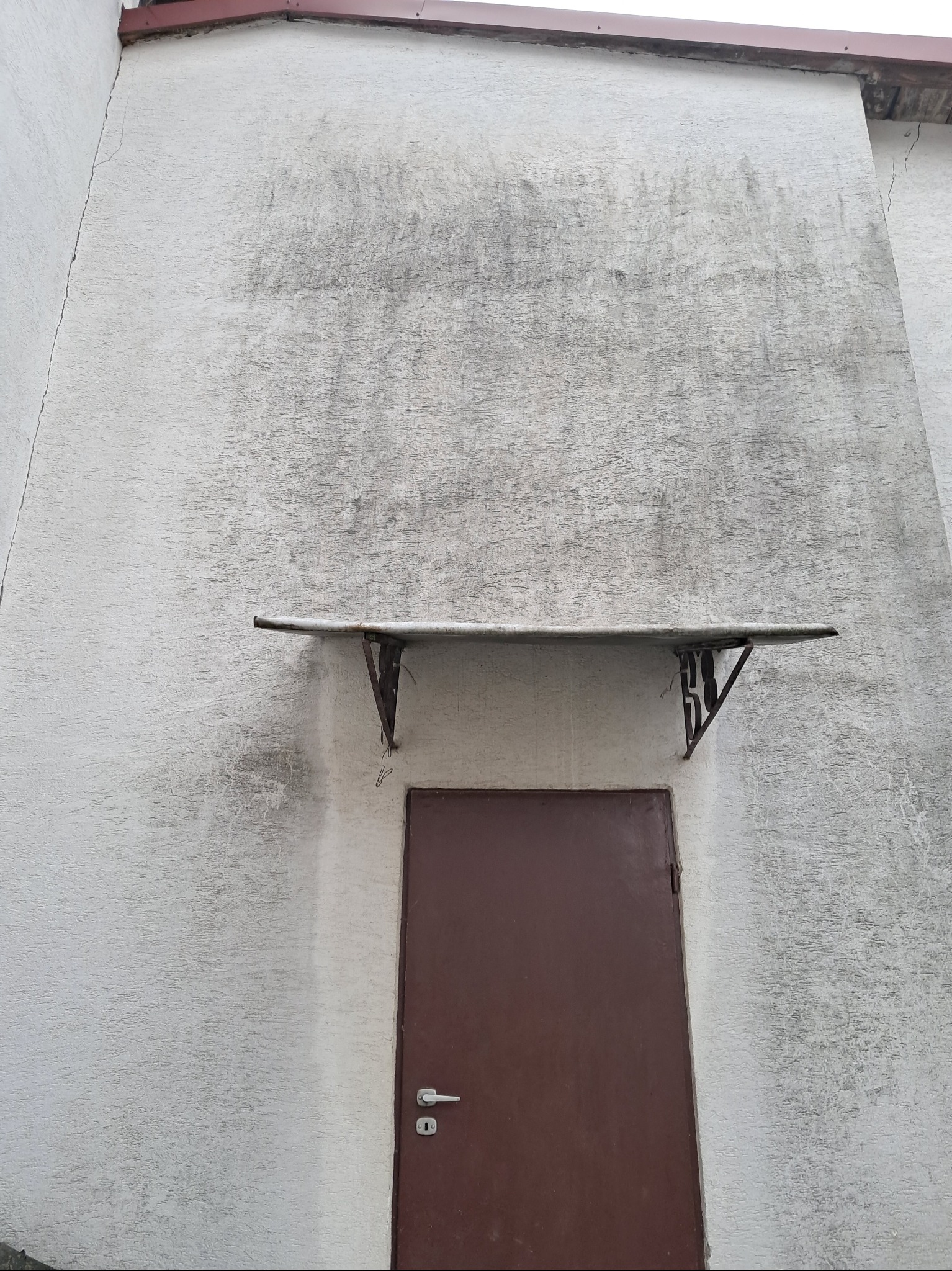 Brudna elewacja budynku w Głowience z brązowymi drzwiami wejściowymi i zadaszeniem. Widoczne zacieki i zabrudzenia na szarej, chropowatej strukturze ściany.