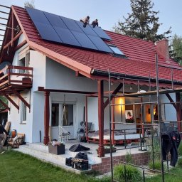 Photon energy Michał Kulik - Montaż paneli fotowoltaicznych na dachu domu jednorodzinnego z czerwoną dachówką, widoczni dwaj monterzy na dachu, w tle ogród i drzewa.