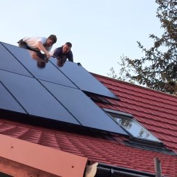 Photon energy Michał Kulik - Dwóch monterów instaluje panele słoneczne na czerwonym dachu domu jednorodzinnego, widoczne okno dachowe i świerk w tle.