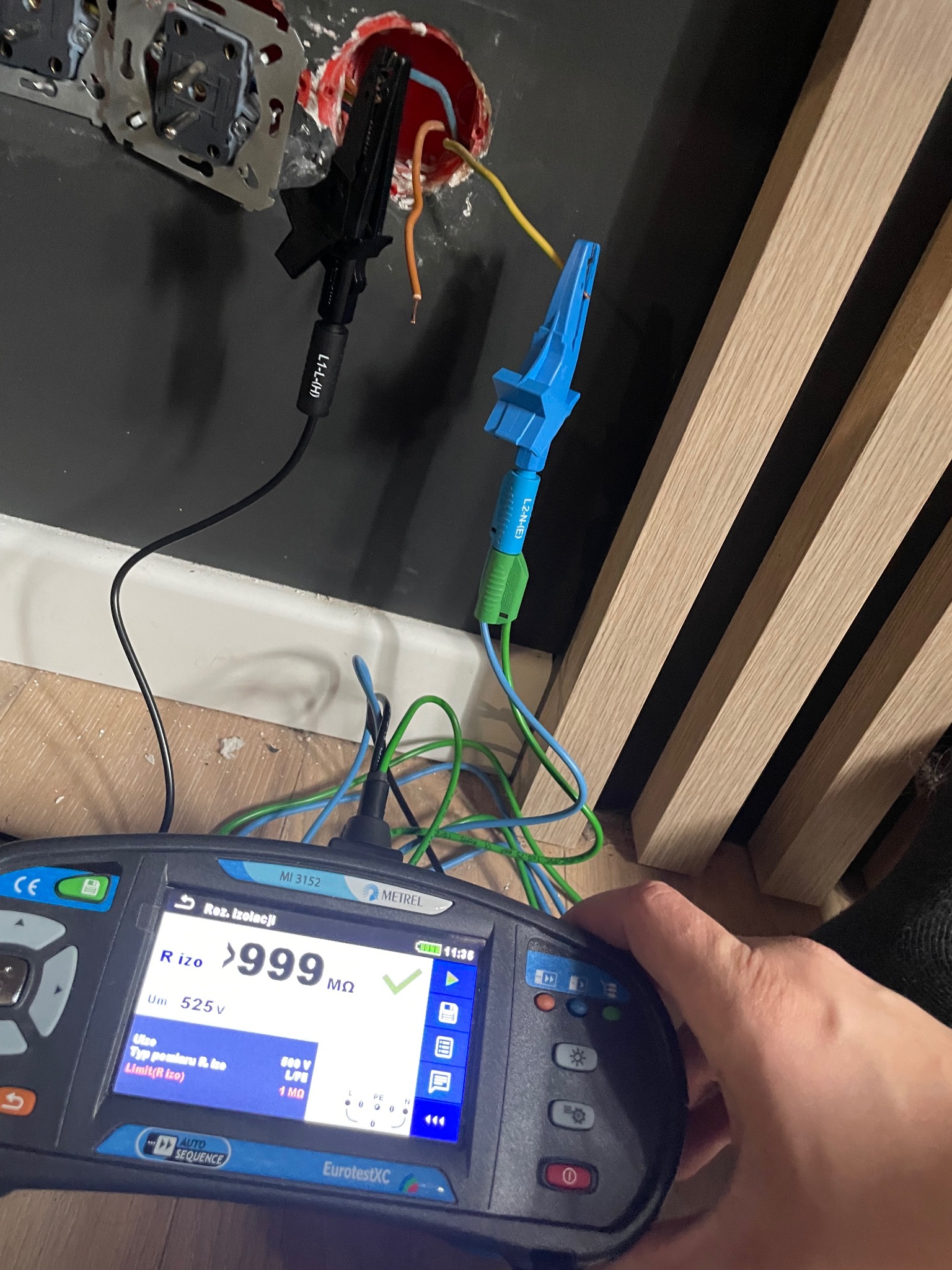 Miernik rezystancji izolacji Metrel MI 3152 z sondami pomiarowymi podłączonymi do przewodów w puszce elektrycznej, wskazujący wynik powyżej 999 MΩ, obok widoczne gniazdka elektryczne i fragment...