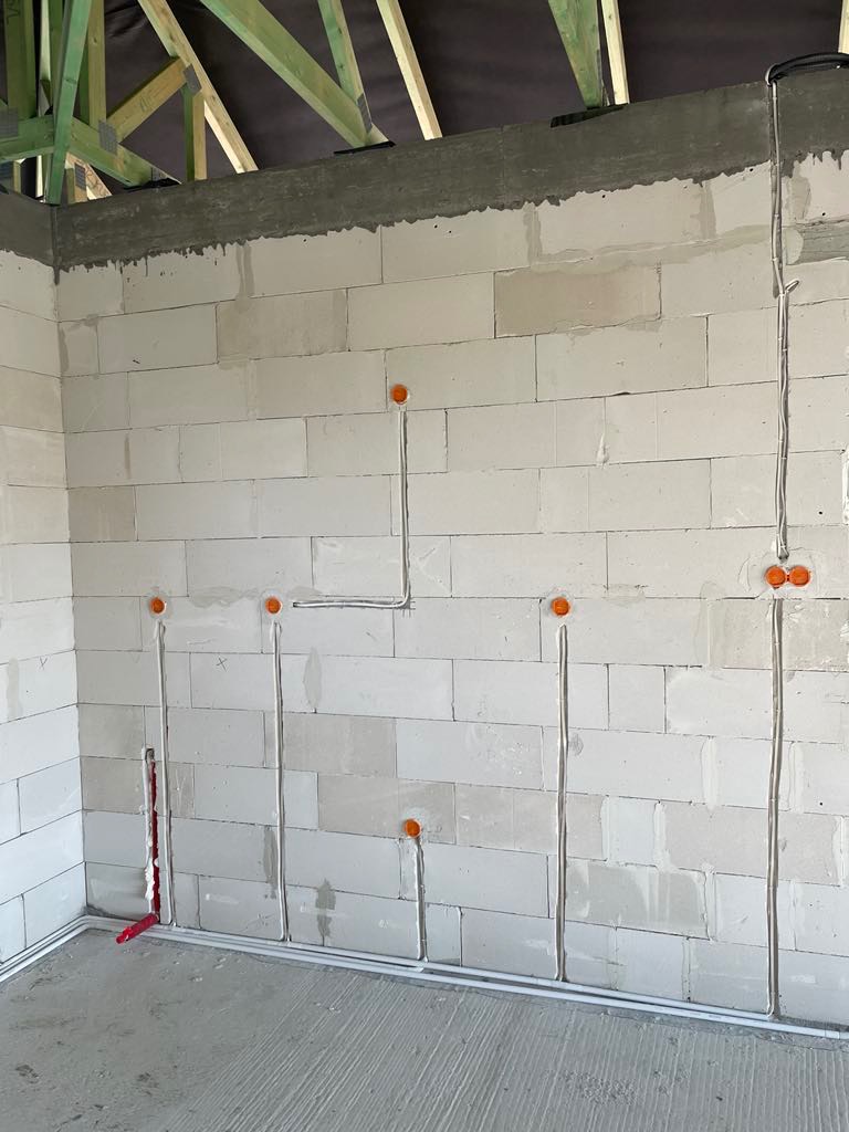 Surowa ściana z betonu komórkowego z widocznymi przewodami elektrycznymi w peszlach, pomarańczowe puszki instalacyjne, widoczne elementy konstrukcji dachu.