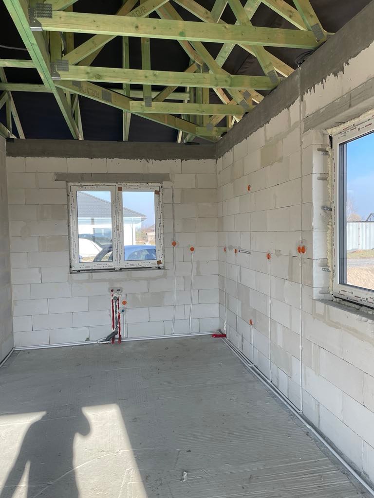 Wnętrze surowego domu w trakcie prac instalacyjnych: widoczne ściany z betonu komórkowego, drewniana konstrukcja dachu, okna PCV oraz rozprowadzone przewody elektryczne i rury instalacji wodnej.