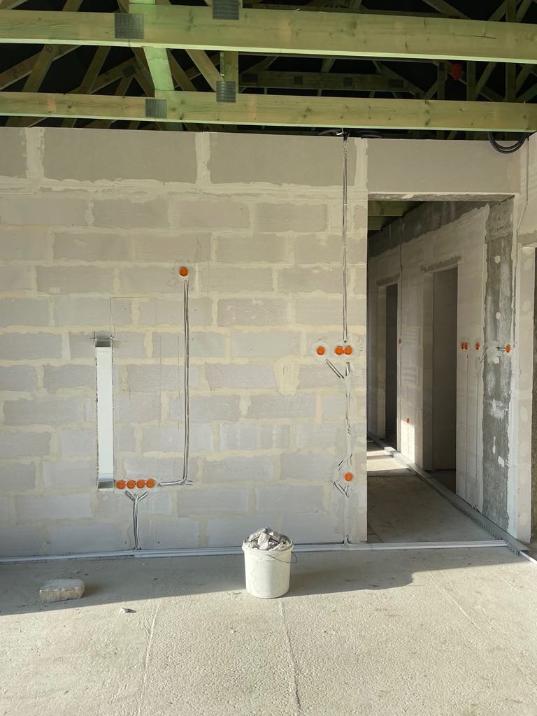 Surowy stan wnętrza domu w trakcie budowy z widocznymi przewodami elektrycznymi, puszkami instalacyjnymi i wiaderkiem gruzu na podłodze.