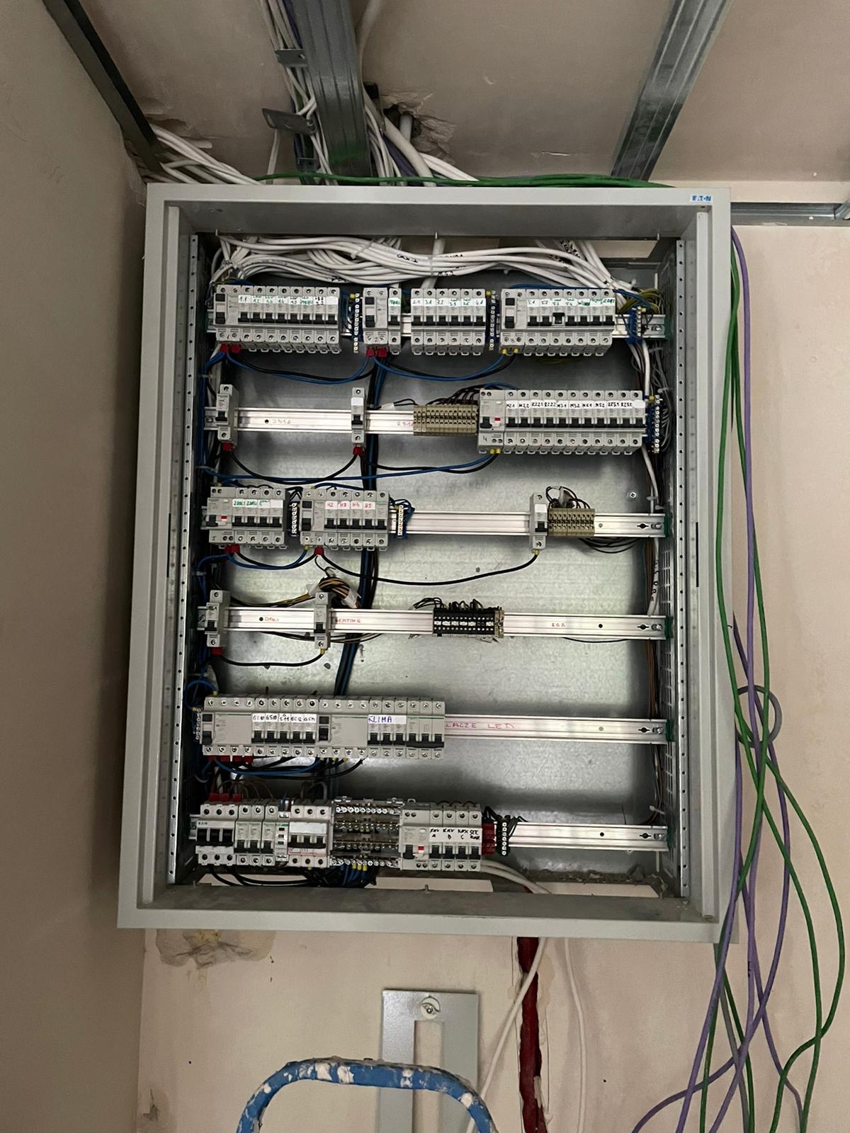 Otwarta skrzynka elektryczna z widocznymi bezpiecznikami, przewodami i listwami montażowymi w surowym wnętrzu.