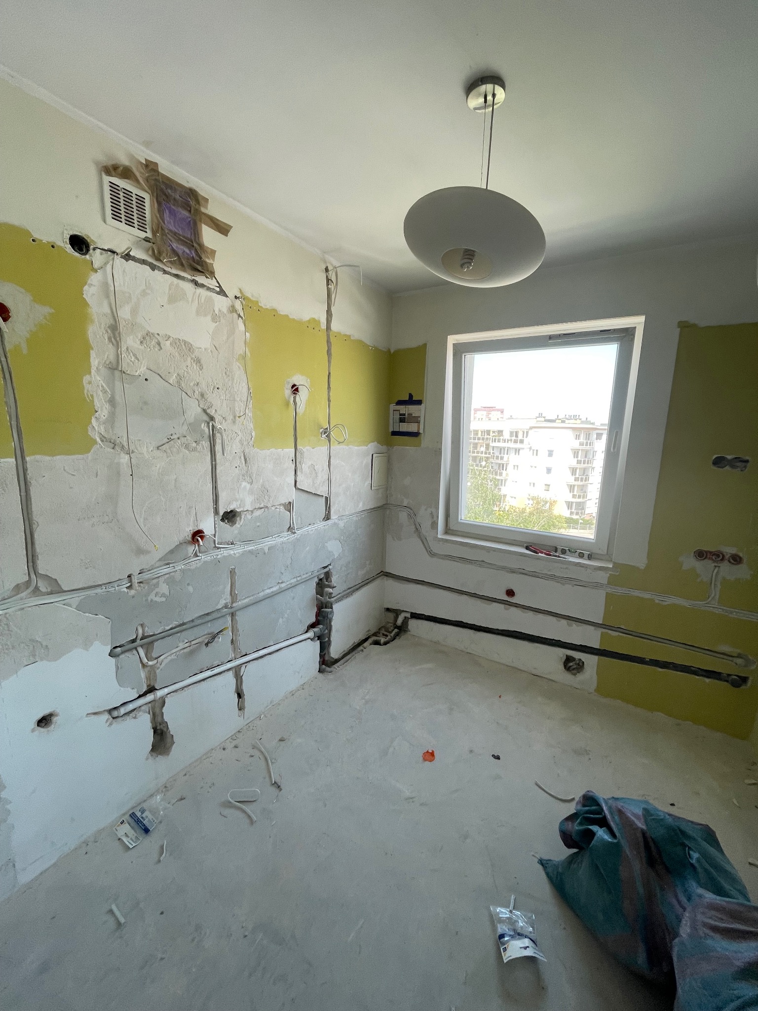 Wnętrze remontowanego pomieszczenia z widocznymi przewodami elektrycznymi i rurami w ścianach, fragmentami odsłoniętej, żółtej farby i prowizorycznie zaklejonym otworem wentylacyjnym.