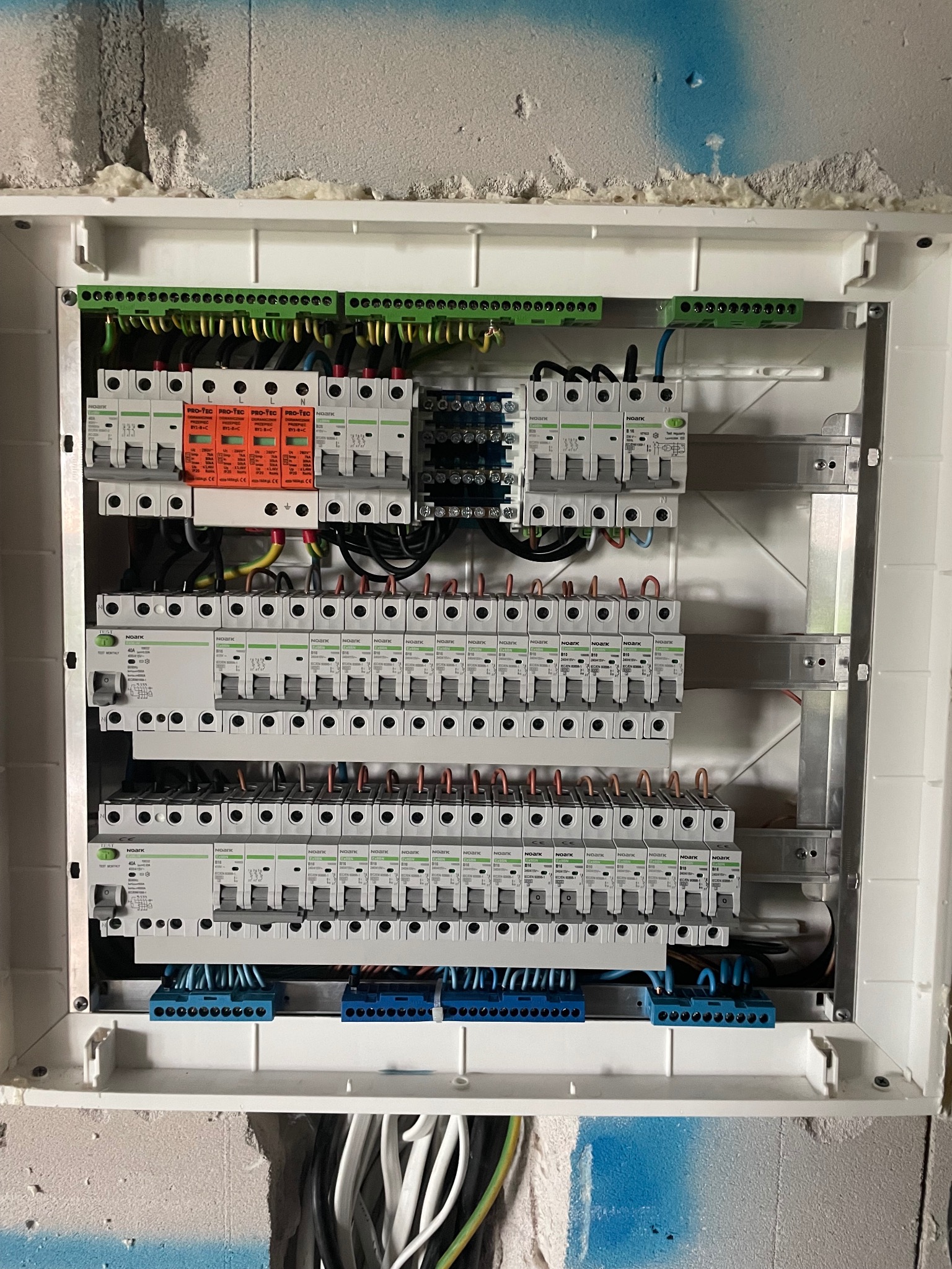 Rozdzielnica elektryczna NOARK z zabezpieczeniami przepięciowymi PRO-TEC, widoczne listwy zaciskowe, przewody i aparatura modułowa, zamontowana w nowym budynku w trakcie wykończenia.