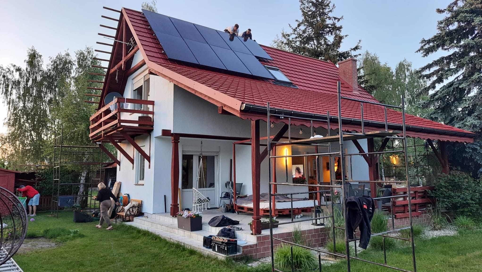 Montaż paneli fotowoltaicznych na dachu domu jednorodzinnego z czerwoną dachówką, widoczni dwaj monterzy na dachu, w tle ogród i drzewa.