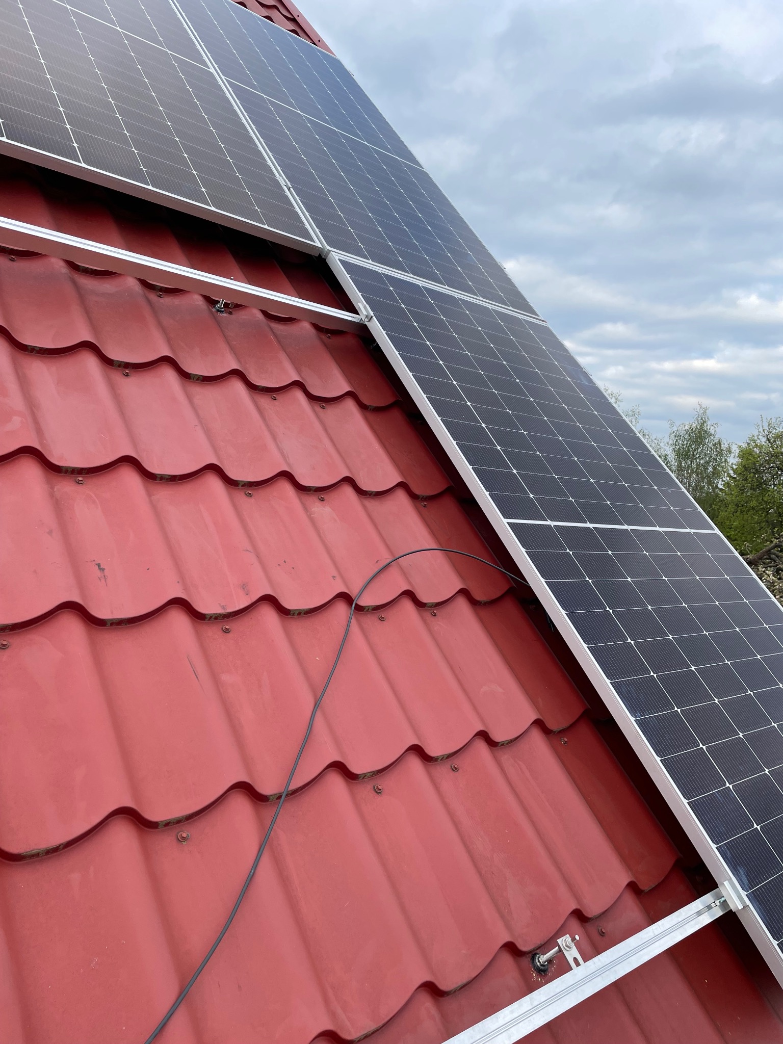 Panele słoneczne zamontowane na czerwonym dachu z dachówki ceramicznej, widoczne mocowania i kabel, pochmurne niebo w tle.