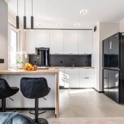 msfoto Marcin Skibiński - Nowoczesna kuchnia z białymi szafkami, czarnym panelem nad blatem, drewnianym blatem wyspowym i czarną lodówką side-by-side; na blacie owoce w misce, nad blatem trzy czarne lampy wiszące.