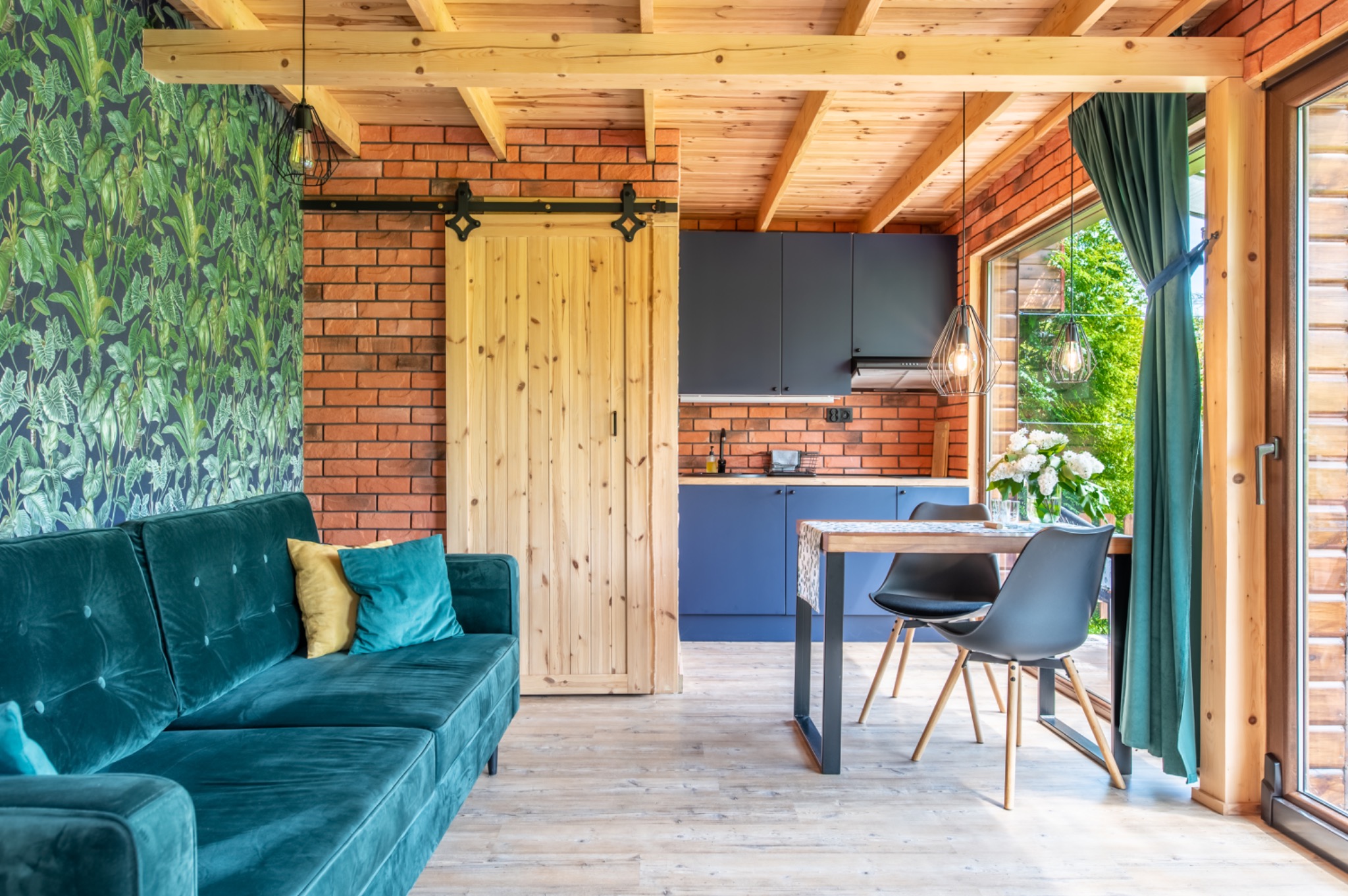 Wnętrze domu z turkusową sofą, ścianą w tropikalne liście, ceglaną ścianą, drewnianymi drzwiami przesuwnymi i kuchnią w tle.