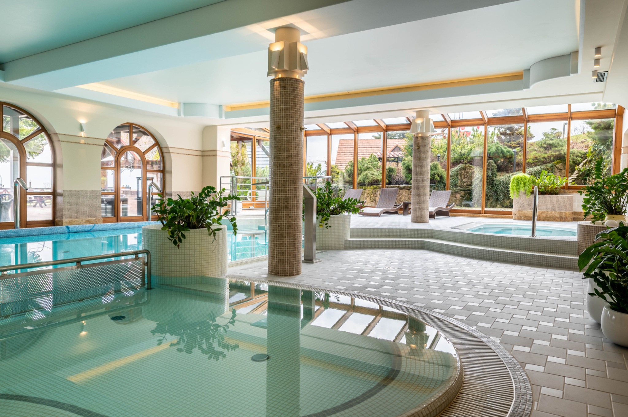 Wnętrze luksusowego spa z basenem, jacuzzi i strefą relaksu z leżakami, widok przez duże okna na zieleń ogrodu.