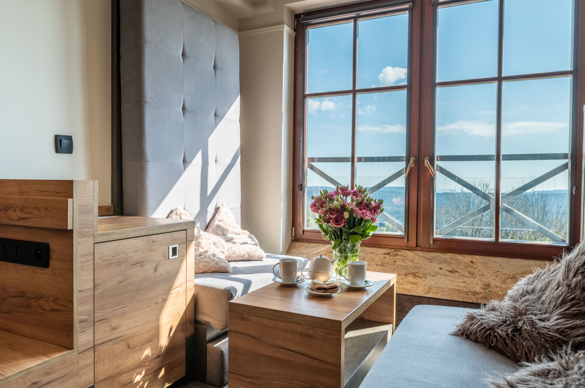 Jasne wnętrze pokoju hotelowego z widokiem na krajobraz przez okno, bukiet różowych kwiatów w wazonie na drewnianym stoliku, dwie filiżanki i czajniczek obok talerzyka z ciastkami.