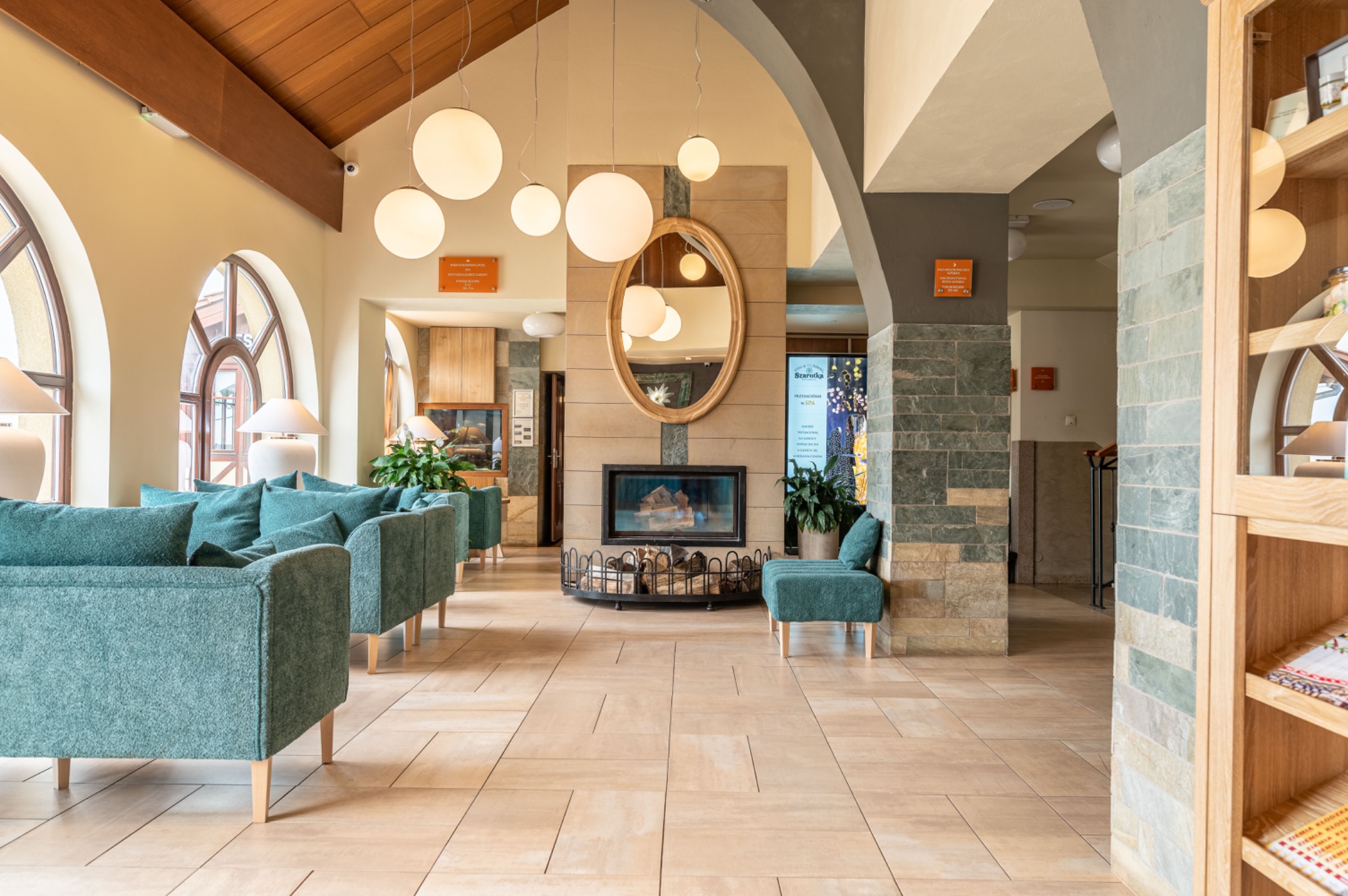 Przestronne lobby hotelowe z turkusowymi sofami, kominkiem z lustrem w ozdobnej ramie i kulistymi lampami wiszącymi.