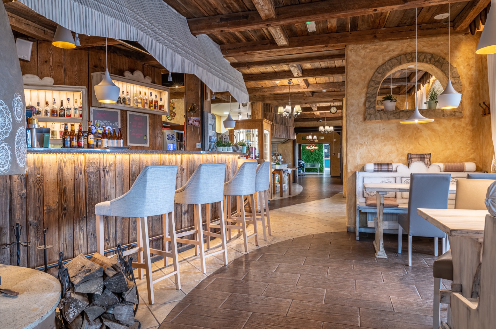 Rustykalne wnętrze baru z drewnianym sufitem i ścianami, oświetlony bar z rzędem krzeseł barowych, widoczne butelki alkoholi, dekoracyjny kominek z drewnem i dalszy plan z miejscami siedzącymi.